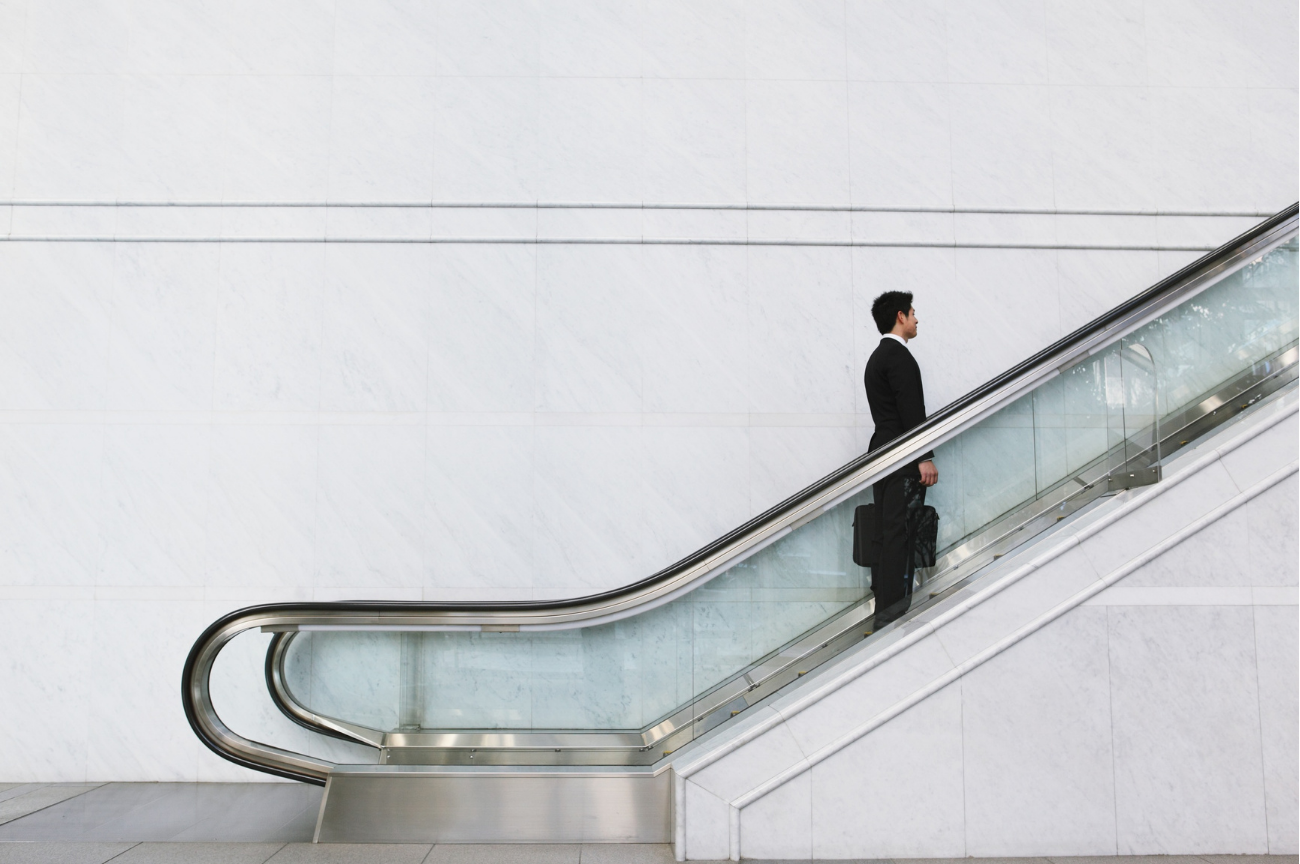 A business professional in a suit carrying a briefcase rides an escalator upward against a clean white marble wall